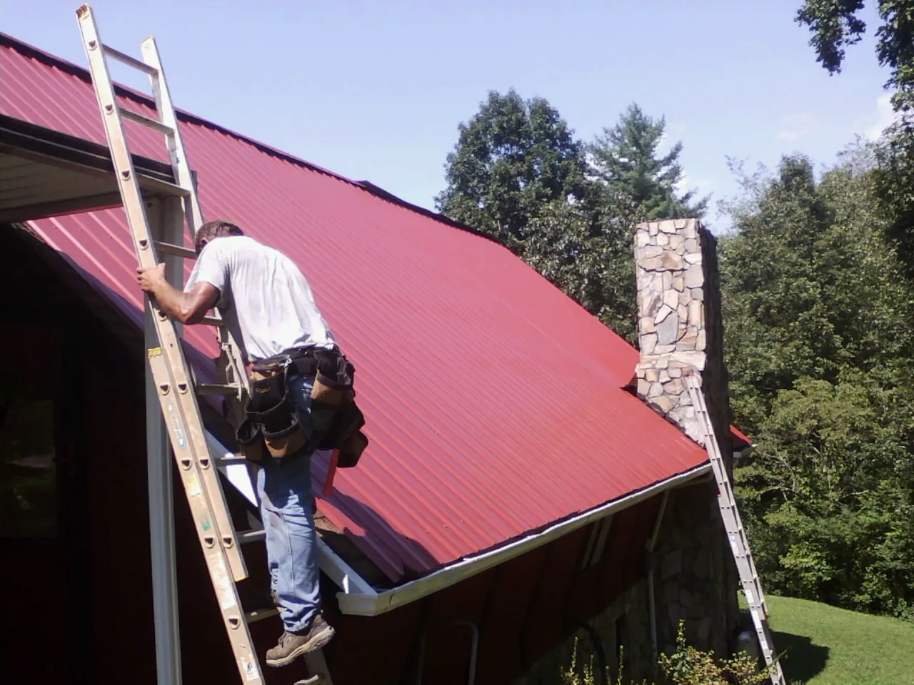 Roofer on a ladder beside a red metal roof and stone chimney — Soffit & Fascia Repair by Roofing Brothers in West Fargo